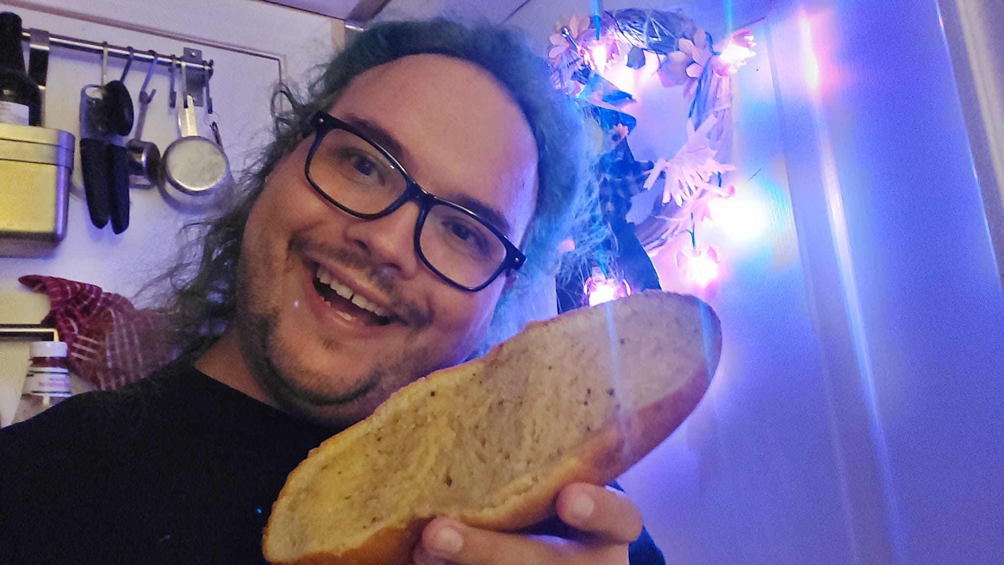 A man with long green hair and no beard holds a piece of garlic bread in a kitchen lit in purple light, Photo 2